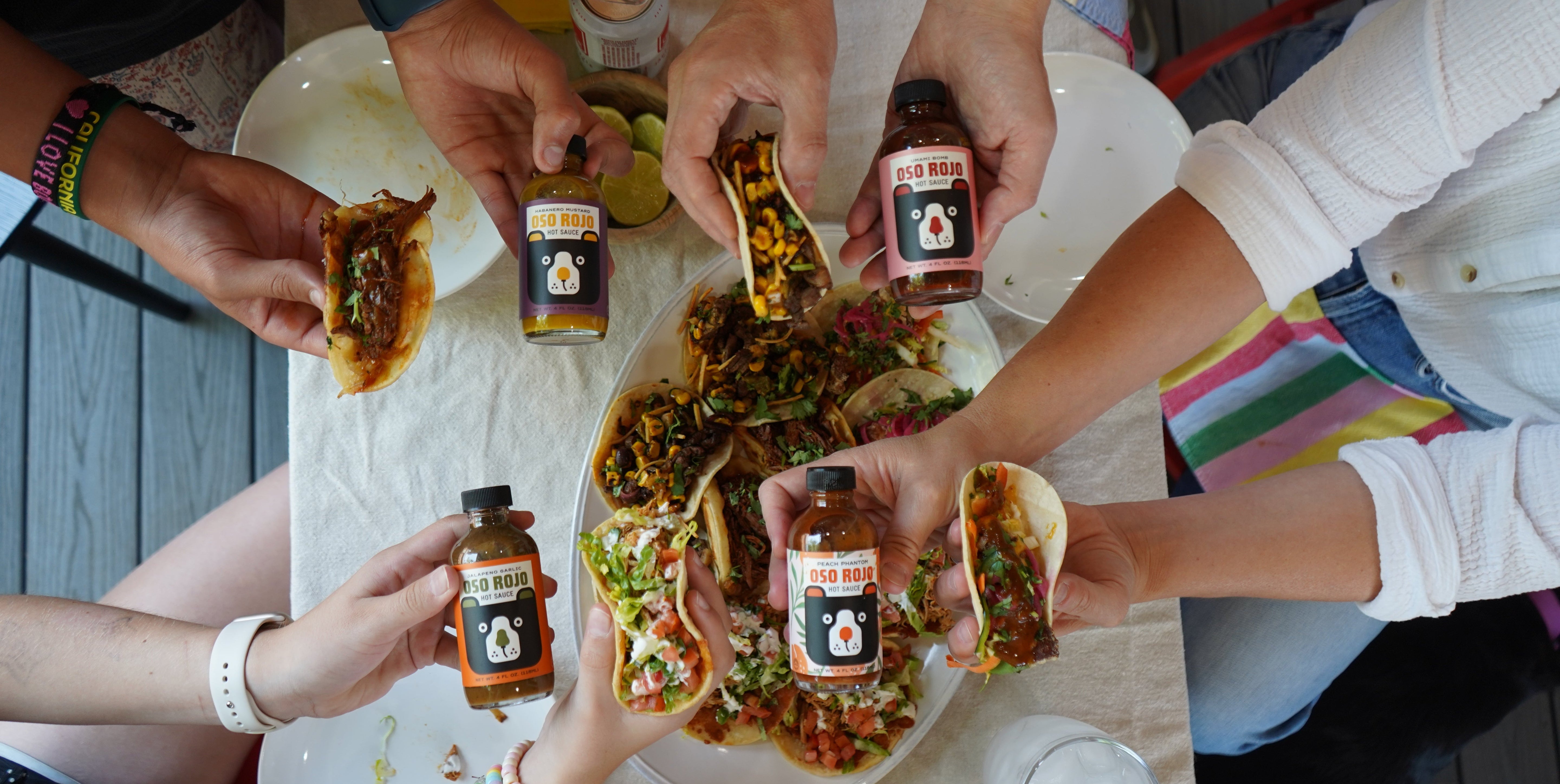 Overhead view of several people reaching toward tacos on a table while holding bottles of hot sauce. The scene is set on a patio with a table covered in food and drinks.