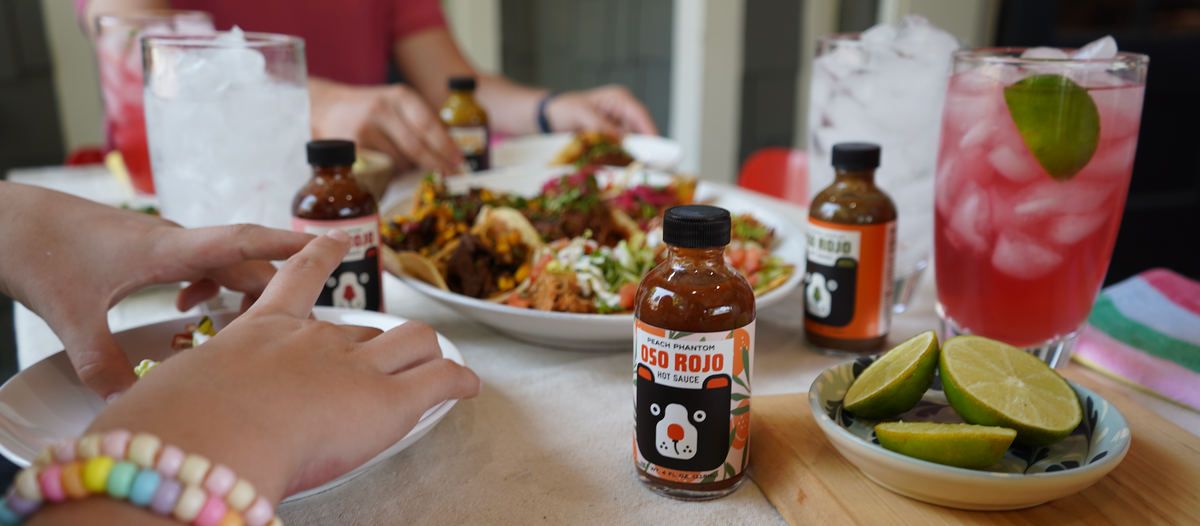 A dining table scene with several bottles of Oso Rojo hot sauce, a plate of tacos, drinks, and visible hands and part of a person seated at the table, so this is Lifestyle rather than Product/Packaging.